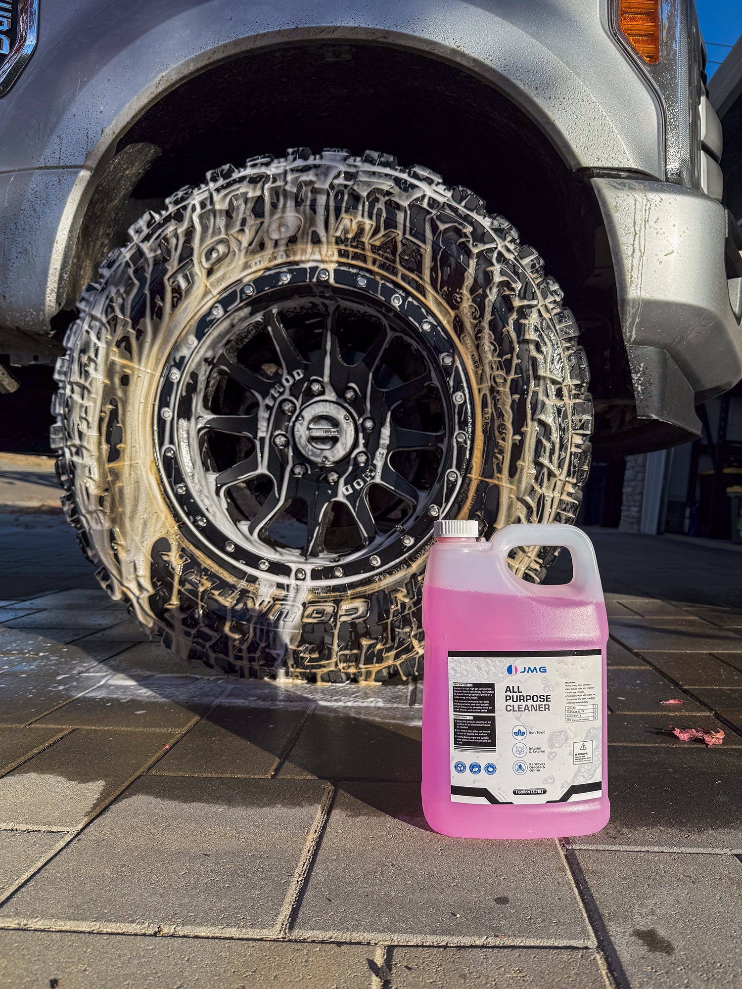 JMG All Purpose Cleaner gallon in front of truck tire and rim covered in foam from IK Foam Sprayer during detailing at San Francisco auto shop
